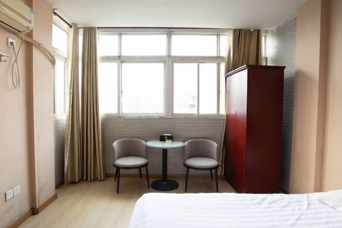 Et hotelværelse med en seng, et rundt bord med to stole, et klædeskab og et stort vindue med gardiner.