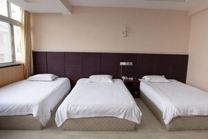 Habitación de hotel con tres camas individuales, cabecera, mesita de noche con lámpara y ventana con cortinas.