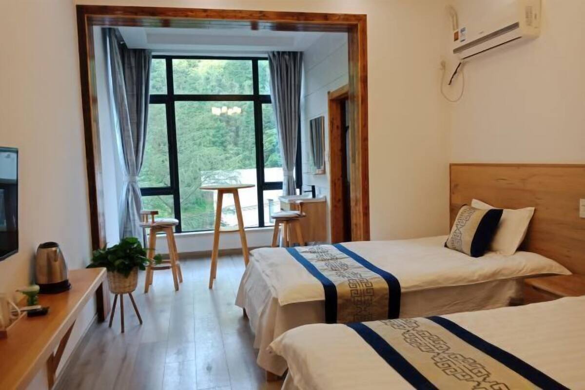 Une chambre d’hôtel avec deux lits, un bureau en bois, une petite table, une plante en pot et une vue sur la verdure grâce à de grandes fenêtres.