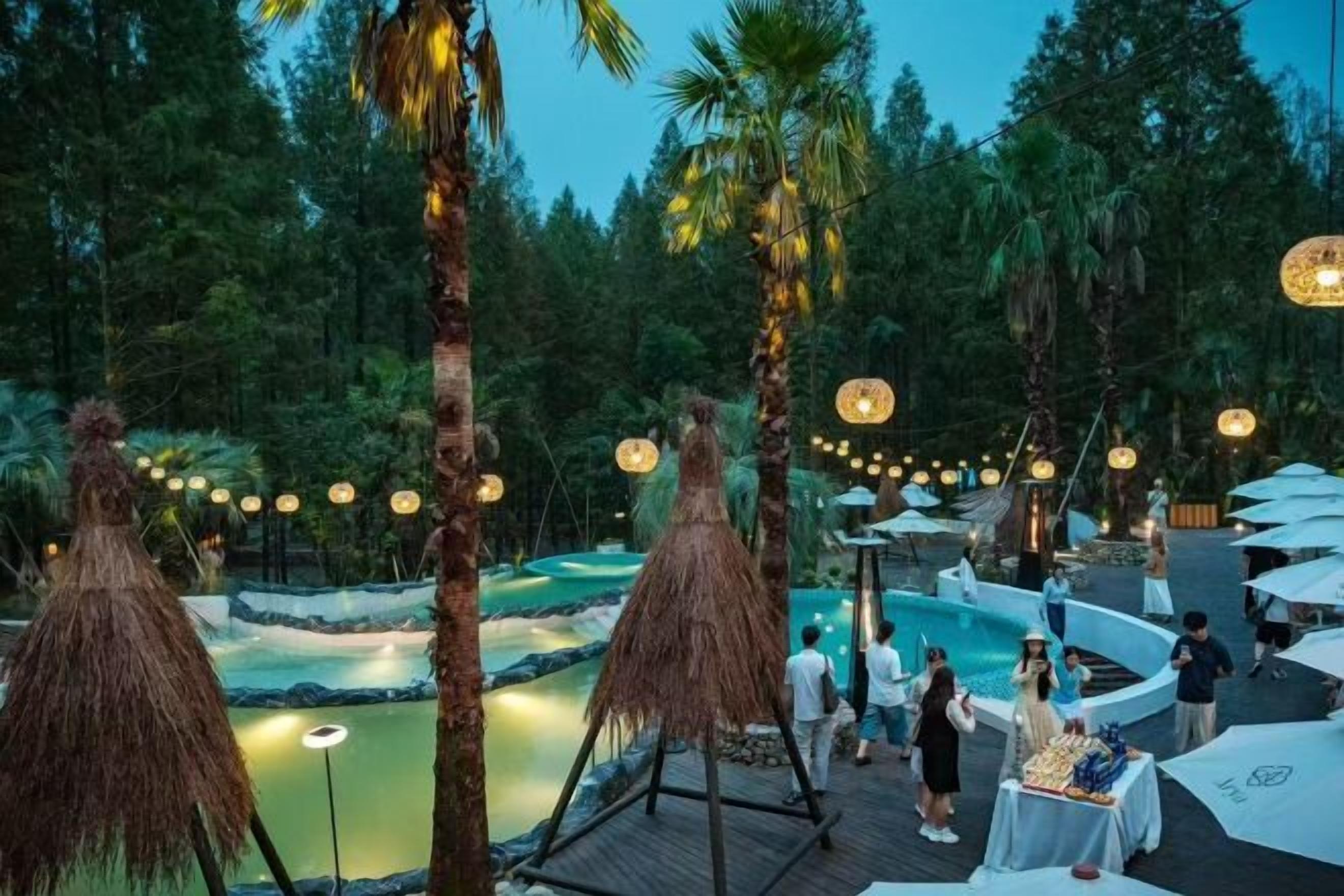 Un área de piscina al aire libre con cubierta de madera, rodeada de palmeras y luces colgantes.