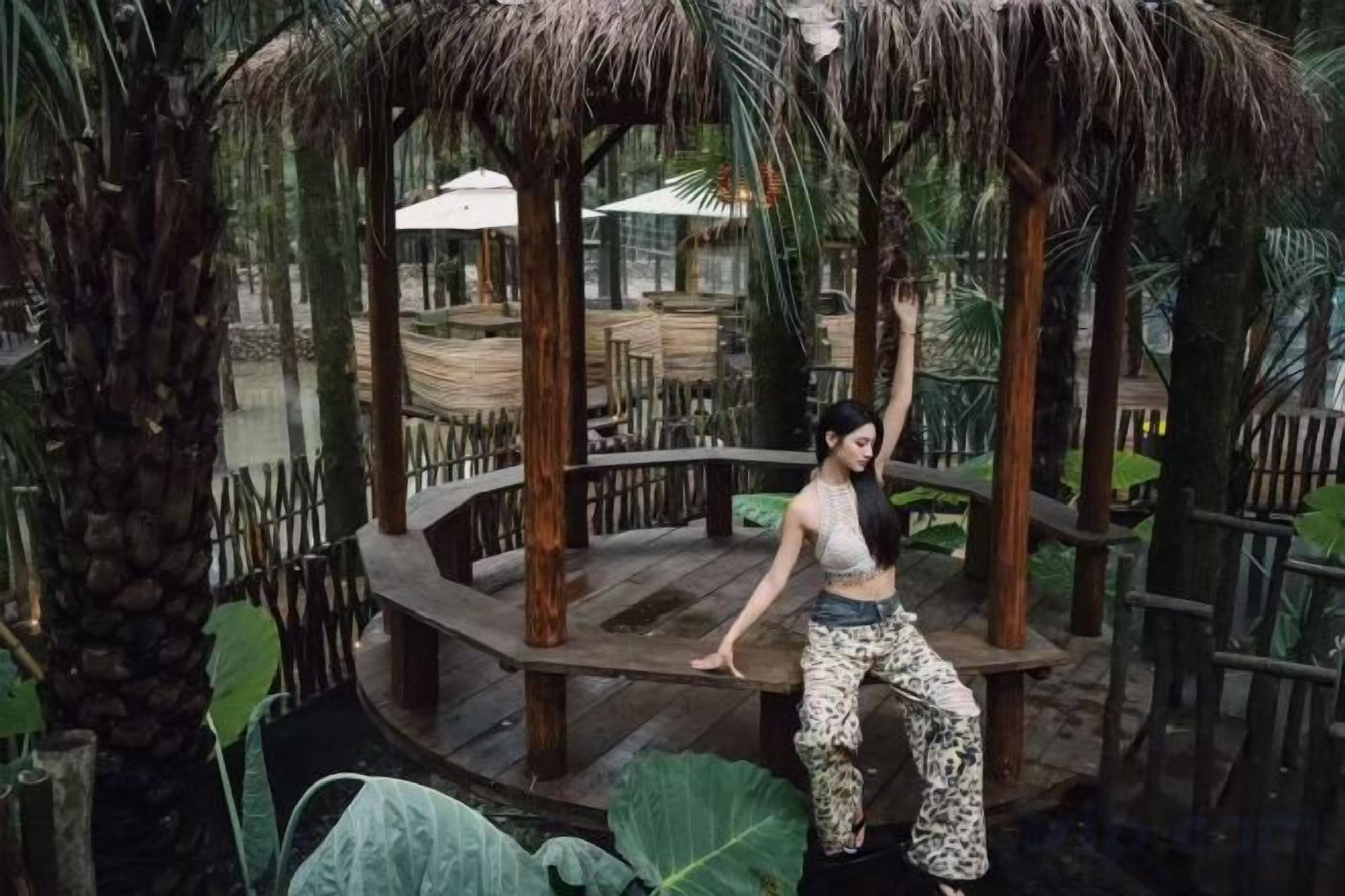 Una persona sobre una plataforma de fusta amb teulada de palla, envoltada de plantes tropicals i una tanca de fusta.