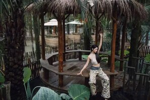 A person standing on a wooden platform with a thatched roof, surrounded by tropical plants and a wooden fence. - Yunxing Villa (Deqing)