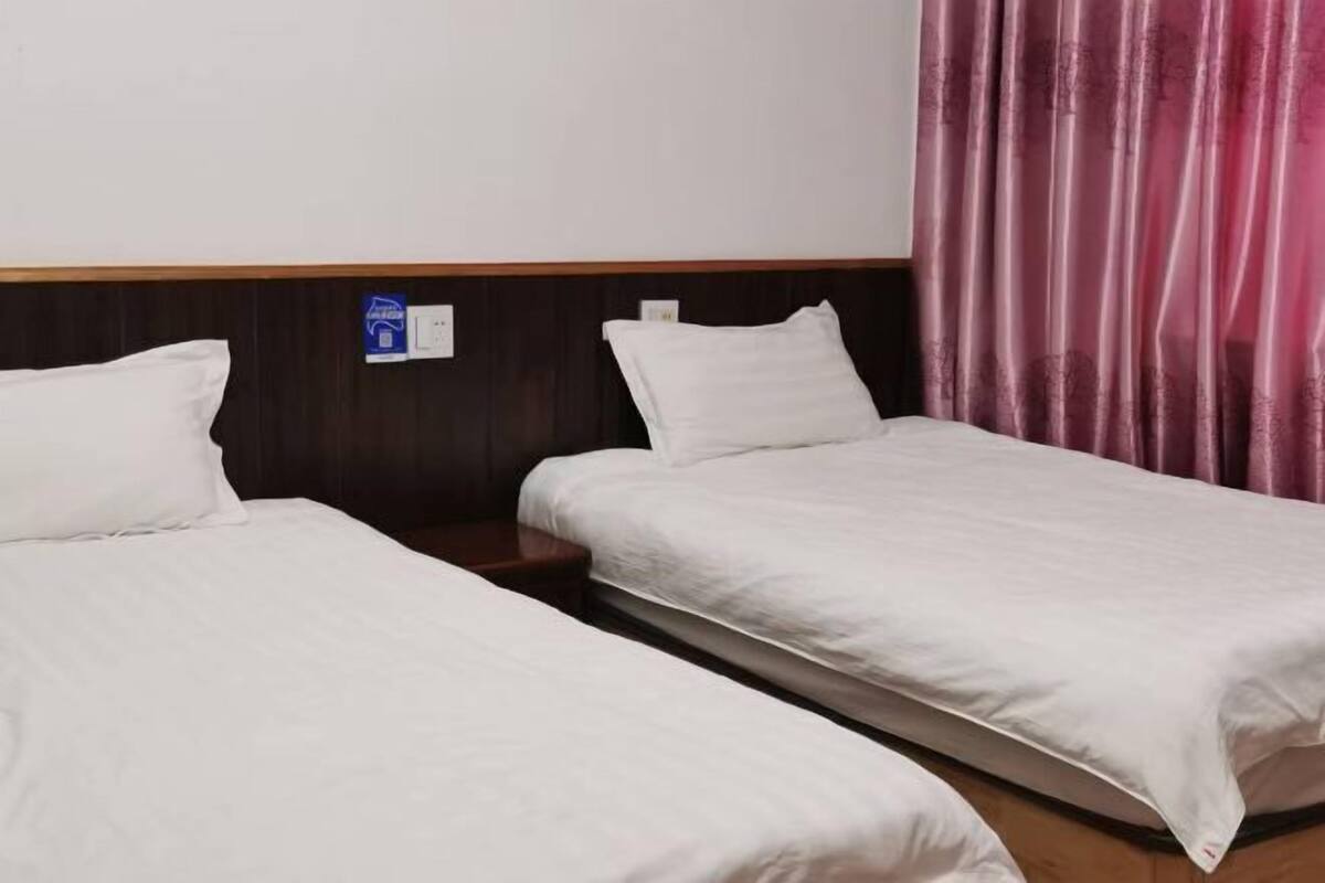 Une chambre d’hôtel avec deux lits simples, une tête de lit en bois, un rideau rose et une prise électrique au mur.