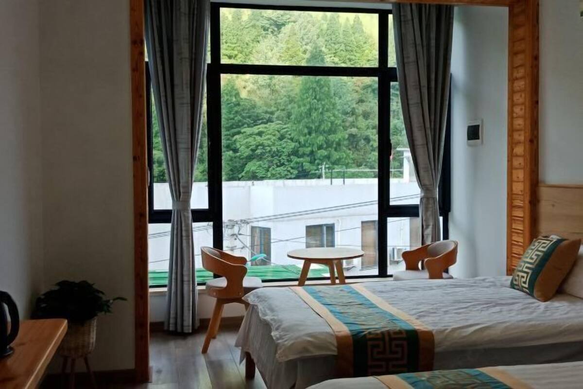 Une chambre d’hôtel avec une grande fenêtre, un lit recouvert d’une courtepointe à motifs, une petite table ronde, deux chaises et un bureau en bois agrémenté d’une plante.