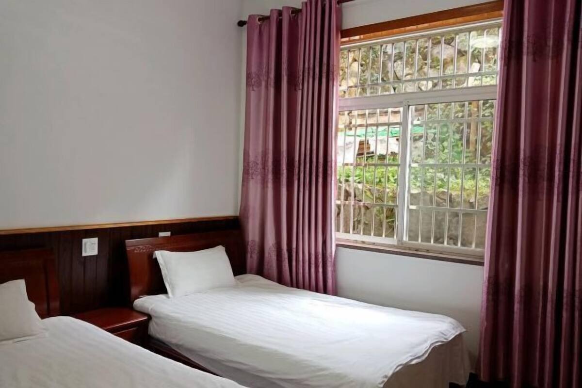Une chambre avec deux lits simples, une tête de lit en bois, une fenêtre avec des barreaux et une vue sur la verdure.