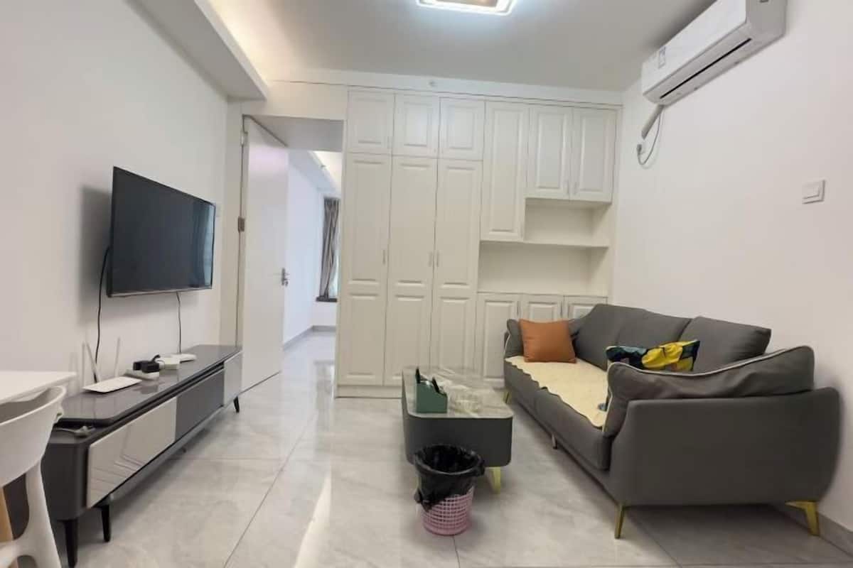 A modern living room with a flat-screen TV, a grey sofa, a coffee table, and a dining area with a white table and chairs.