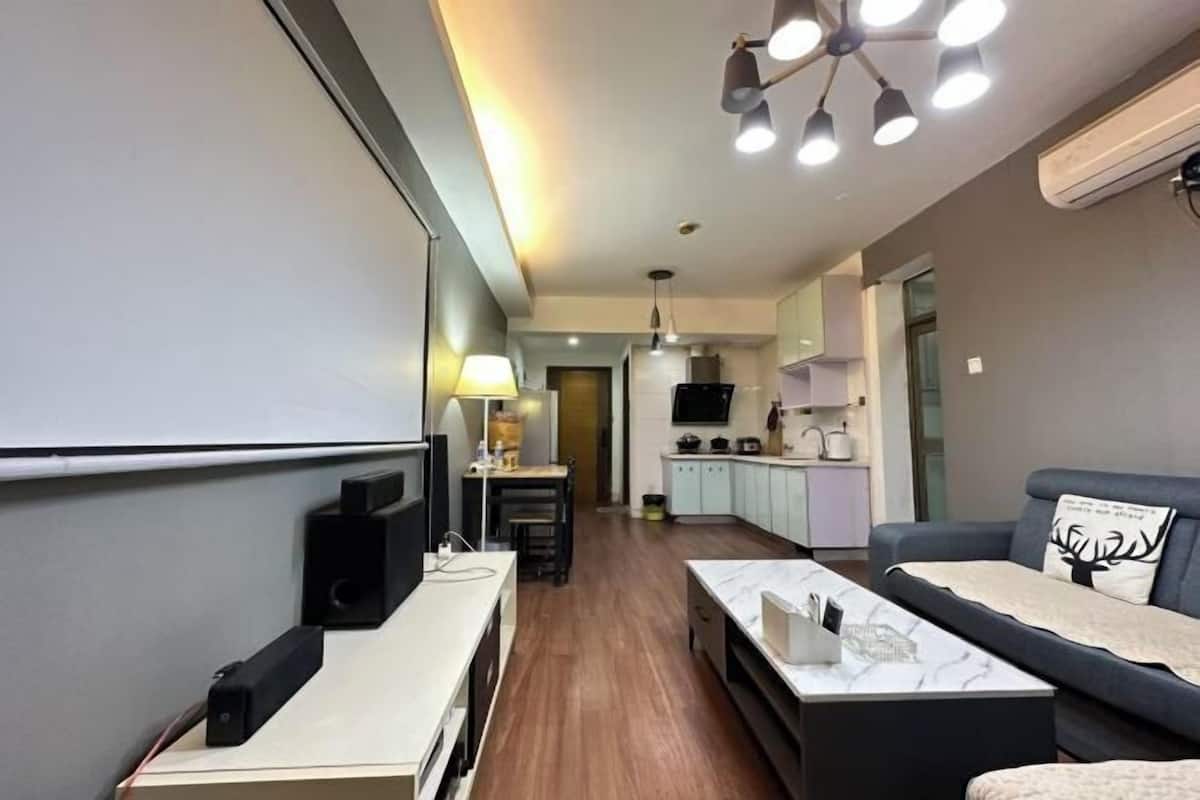 A modern living room with a sofa, a coffee table, and a TV. There is a kitchen area in the background with cabinets and a microwave.