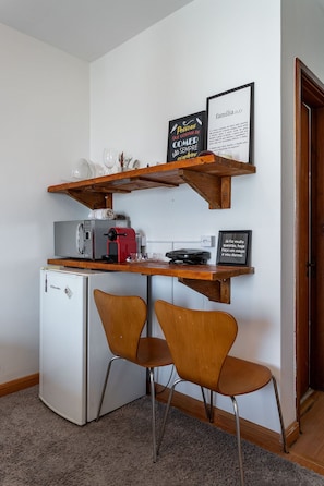 Une cuisine compacte équipée d’un réfrigérateur, d’un four à micro-ondes et d’une machine à café posés sur le plan de travail. On y trouve deux chaises en bois et une étagère au-dessus du plan de travail, où sont rangés des verres et un cadre contenant une citation.