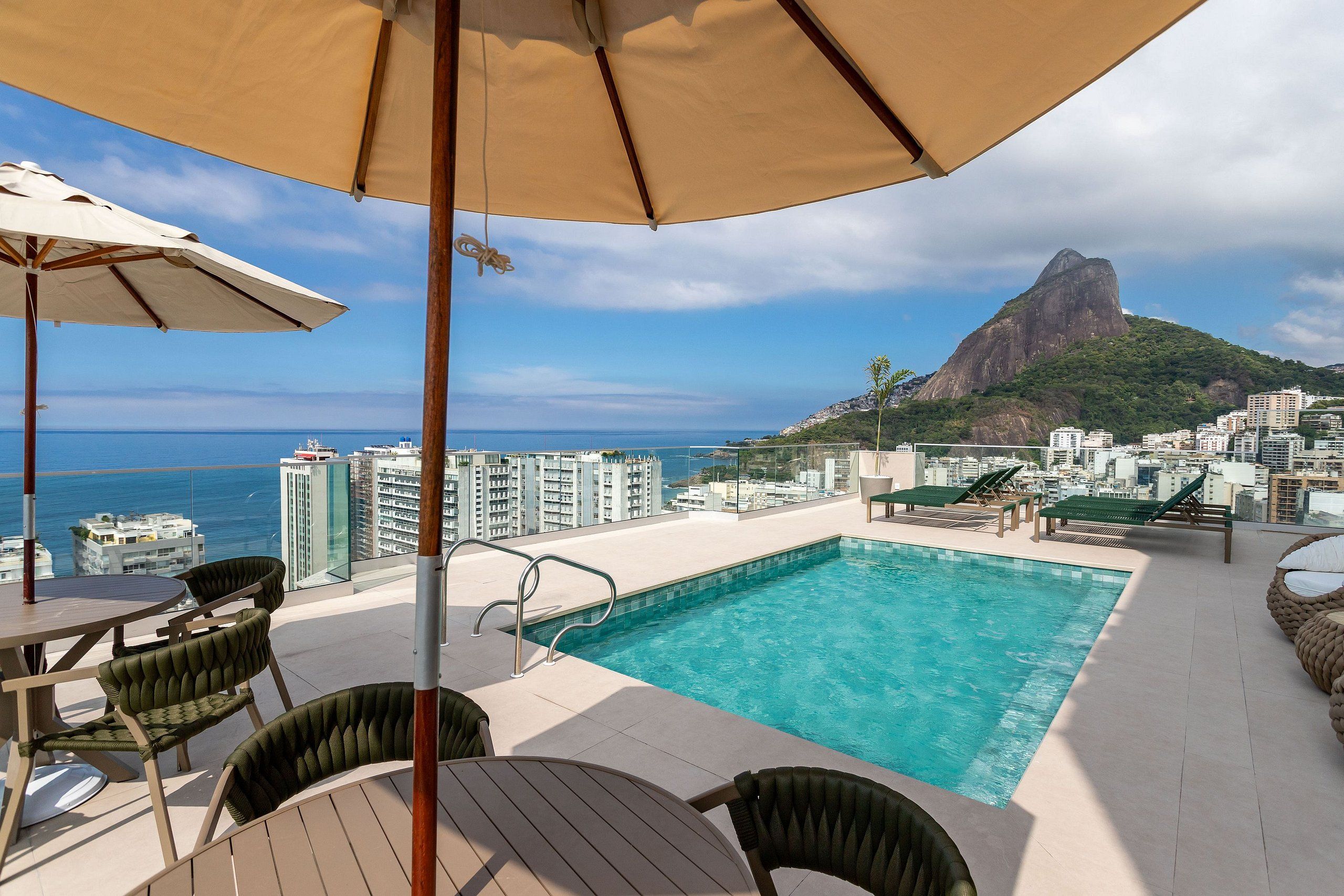 Una piscina al aire libre con sillones de descanso y sombrillas, con vistas a un paisaje urbano y una montaña destacada.