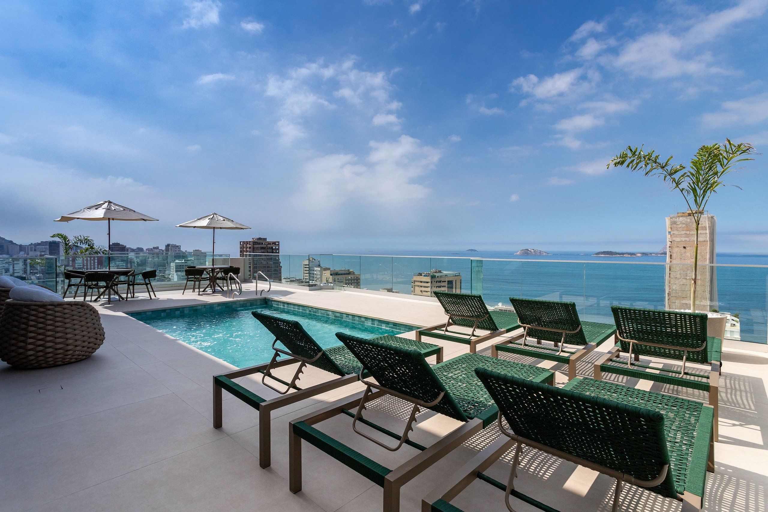 Un área de piscina en la azotea con sillones de descanso y sombrillas, con vistas a un horizonte urbano y al mar.