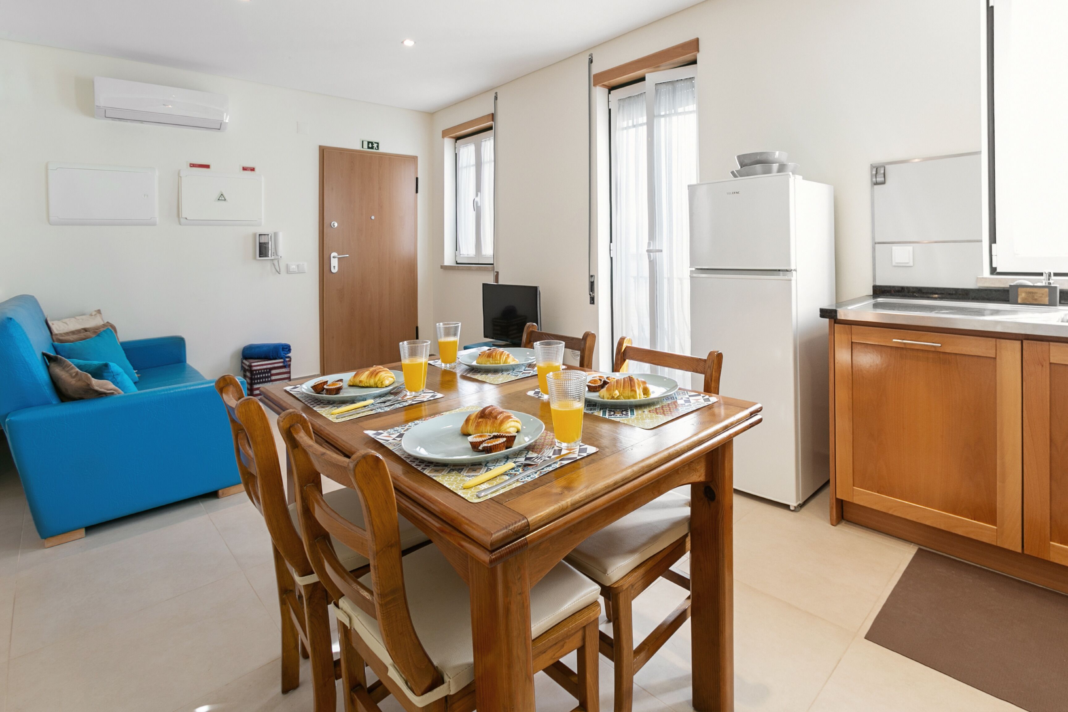 A dining area with a wooden table set for breakfast, a blue sofa, and a kitchen with wooden cabinets and a refrigerator.