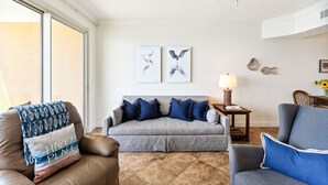 A living room with a sofa, a coffee table, and a side table with a lamp. There are two wall-mounted paintings and a ceiling fan.