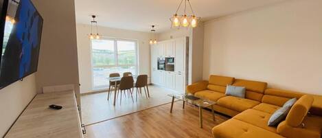 A modern living room with a sofa, a coffee table, and a TV. There is a dining area with chairs and a table. The room has wooden flooring and a large window.