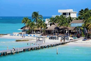 Een hotel aan het strand met een steiger, strandparasols en palmbomen.