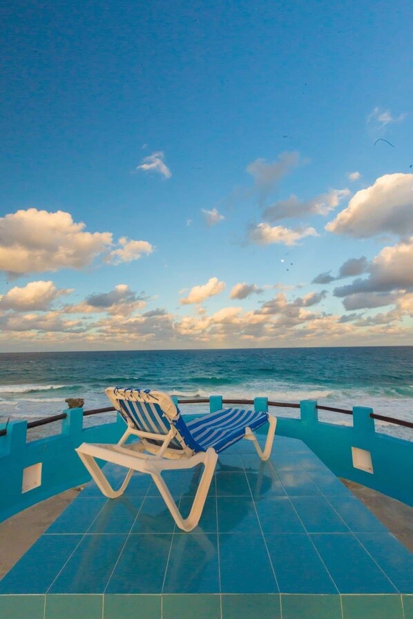 A poolside area with two lounge chairs facing the ocean, a tiled deck, and a railing.