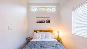 Ein Schlafzimmer mit einem Bett, einem Nachttisch, einer Lampe, einem Fenster und einem Wandbild.