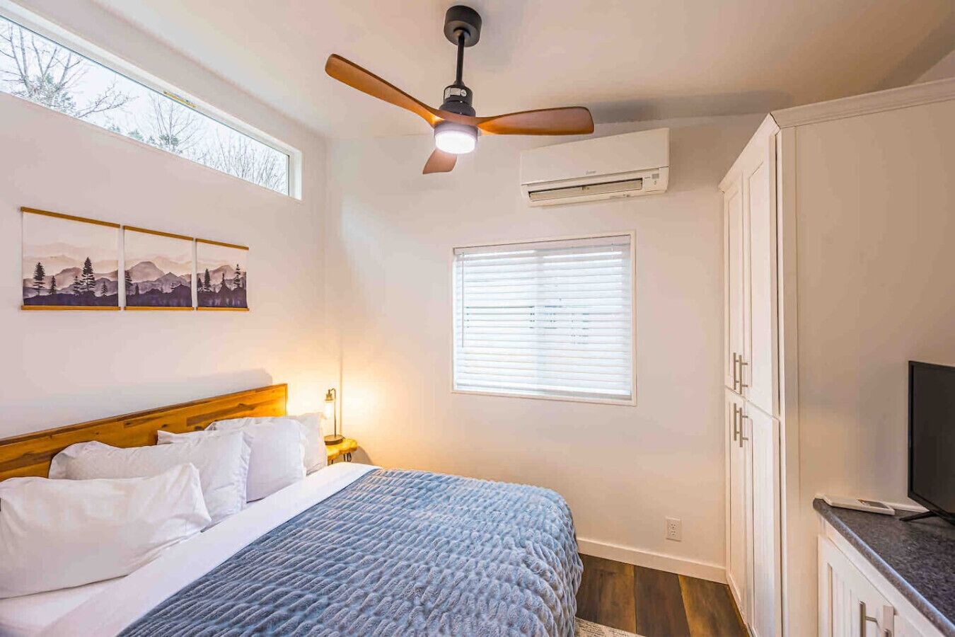 A bedroom with a bed, a ceiling fan, an air conditioner, a window with blinds, and a TV on a cabinet.