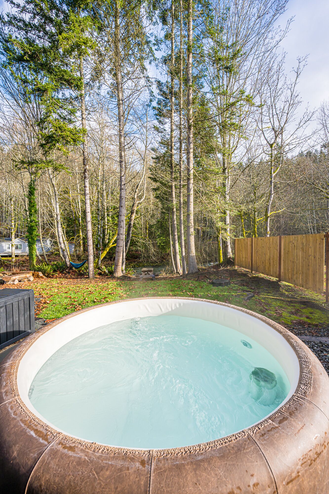 Un jardin avec un jacuzzi entouré d’arbres et délimité par une clôture en bois.