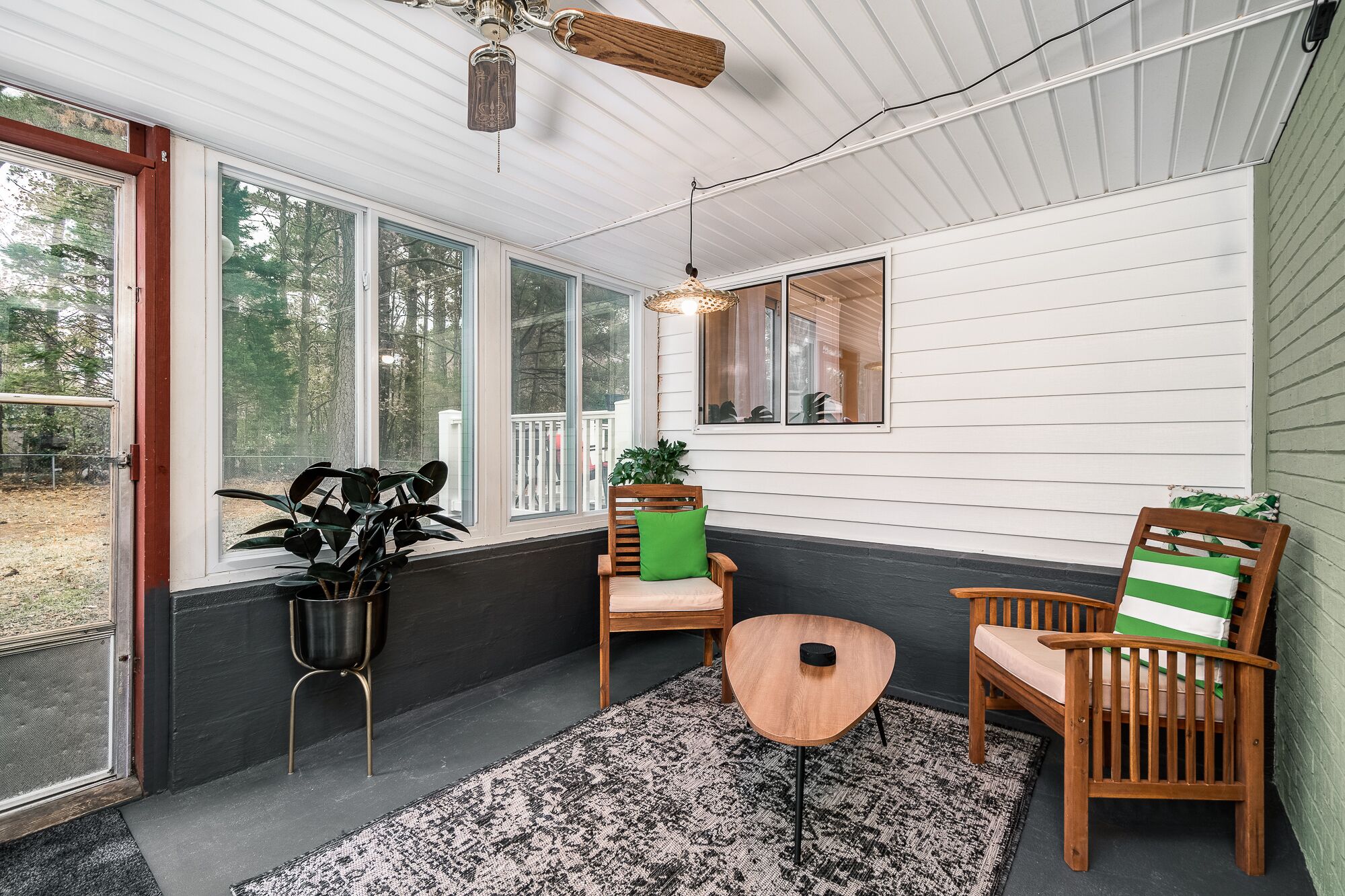 Une véranda avec de grandes fenêtres, un ventilateur de plafond et deux fauteuils en bois avec des coussins.