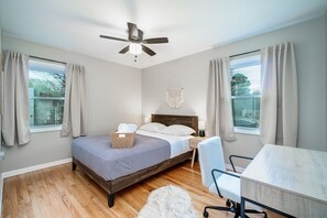 A bedroom with a bed, a desk, a chair, a ceiling fan, and a window with curtains.