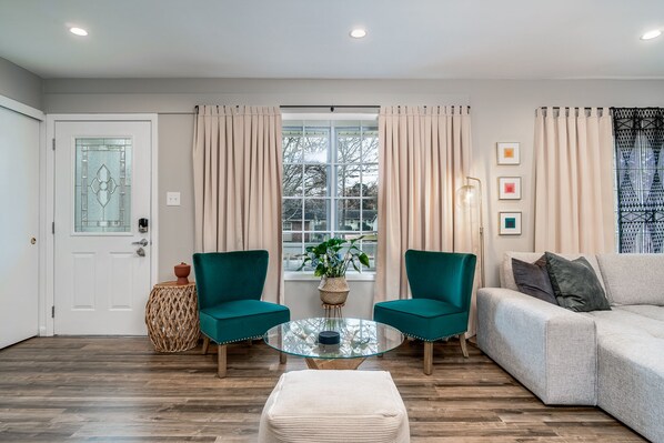 A living room with a glass coffee table, two teal armchairs, a beige sofa, and a white door with a stained glass window.
