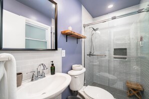 A bathroom with a white sink, a toilet, a glass shower enclosure, and a wooden shelf.