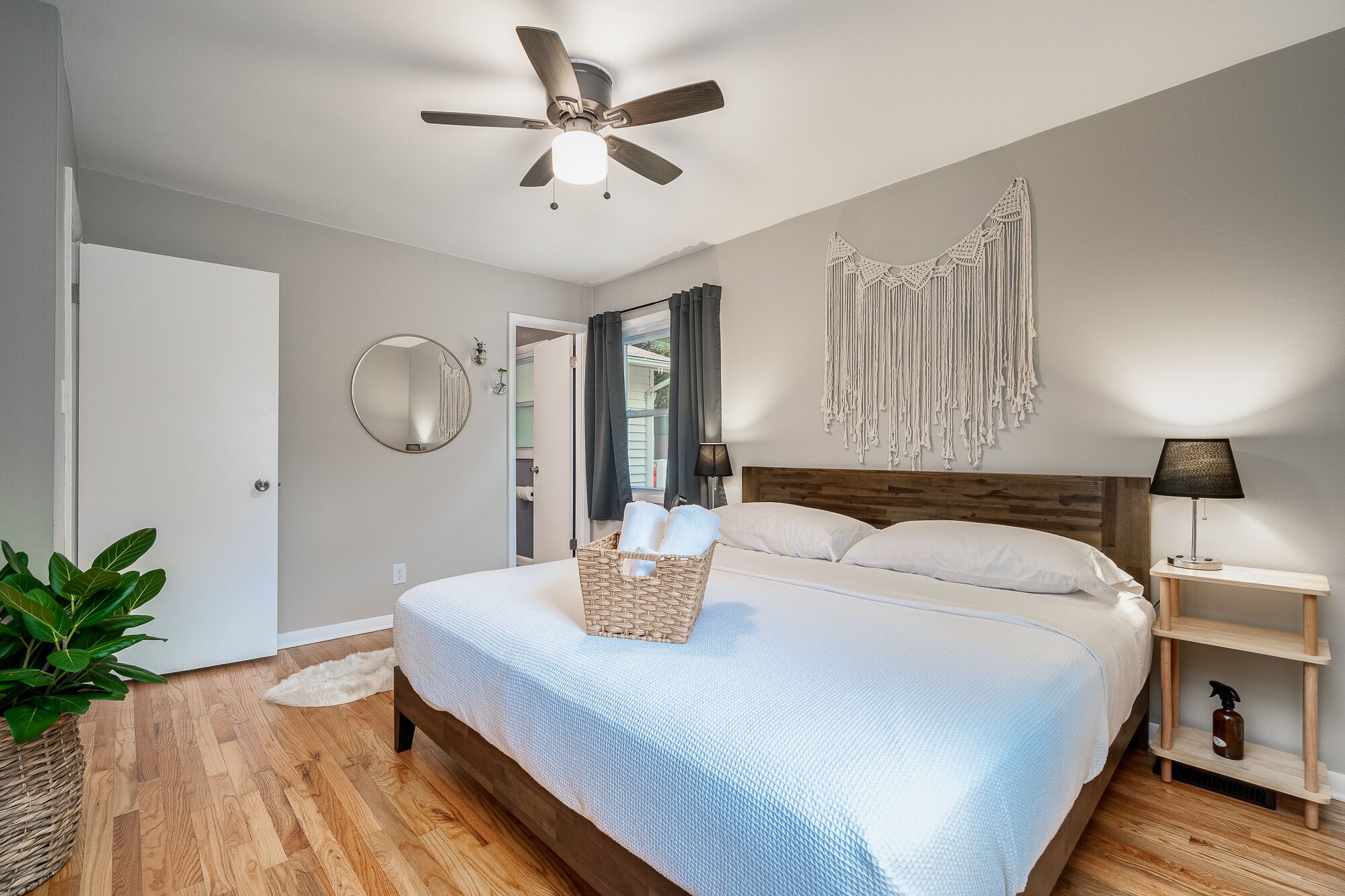 Une chambre avec un lit, un ventilateur de plafond, un miroir fixé au mur, une décoration murale en macramé et une table de chevet avec une lampe.