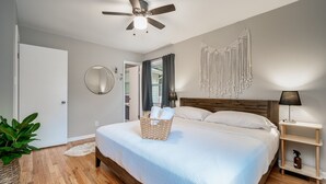 Une chambre avec un lit, un ventilateur de plafond, un miroir fixé au mur, une décoration murale en macramé et une table de chevet avec une lampe.