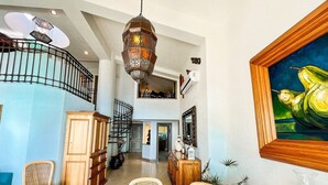 A spacious interior with a high ceiling, a central staircase, wicker furniture, and a large painting.