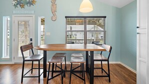 Un comedor con una mesa de madera y cuatro sillas, una lámpara colgante, un adorno en la pared y una ventana con persianas.