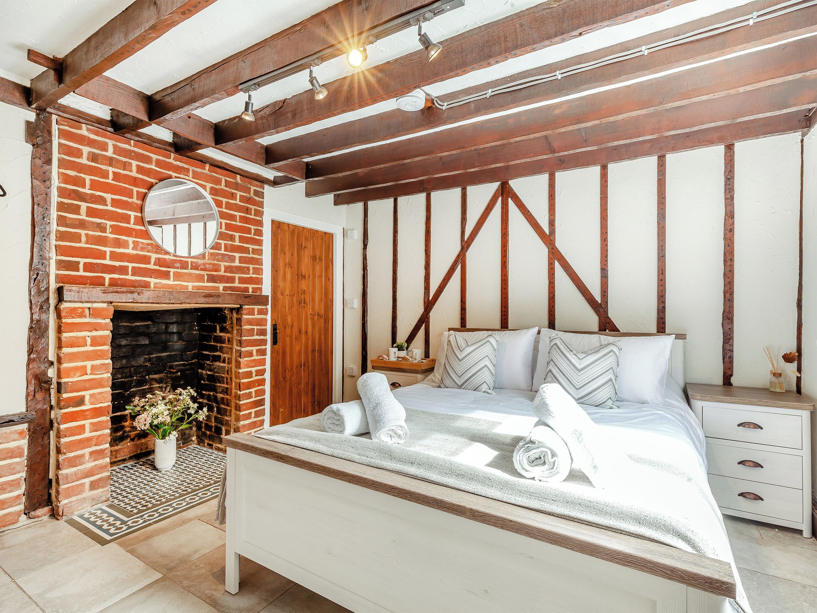 Een slaapkamer met een haard van baksteen, een bed met opgerolde handdoeken, houten balken en een nachtkastje met een lamp.