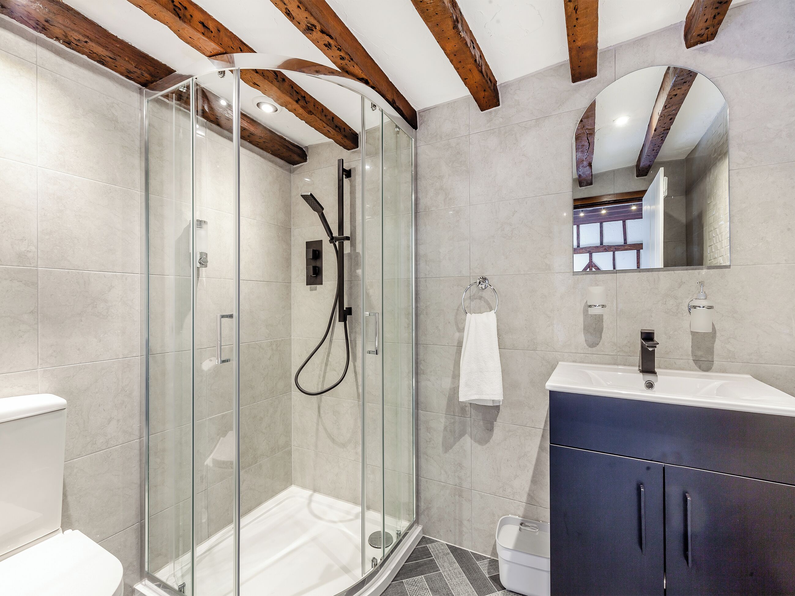 Et moderne badeværelse med en brusekabine med glasdøre, et hvidt toilet, en vaskebord med et mørkt skab og et spejl over vasken.