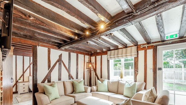 A room with exposed wooden beams, a sectional sofa, a small table, and a window with a view of greenery. - 3 bedroom accommodation in Chelmsford (Chelmsford)