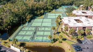 Luftaufnahme eines Tennisplatzkomplexes mit mehreren Plätzen, umgeben von Bäumen und einem Gewässer.