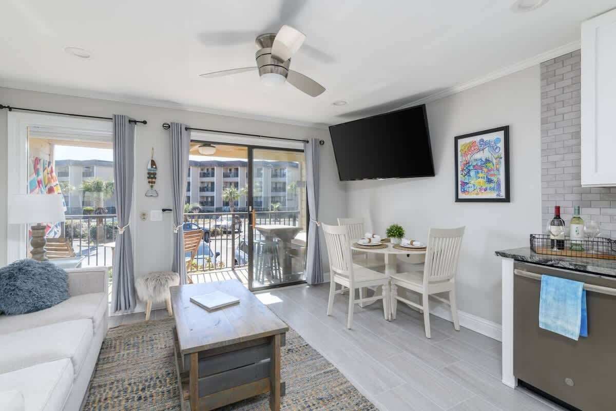 Una sala de estar moderna con sofá, mesa de centro y comedor. Cuenta con un balcón con vistas a edificios y un ventilador de techo.