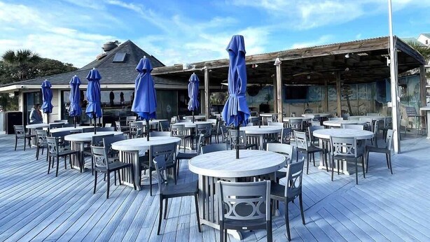 Ein Essbereich im Freien mit runden Tischen und Stühlen, beschattet von blauen Sonnenschirmen, und eine Holzterrasse.