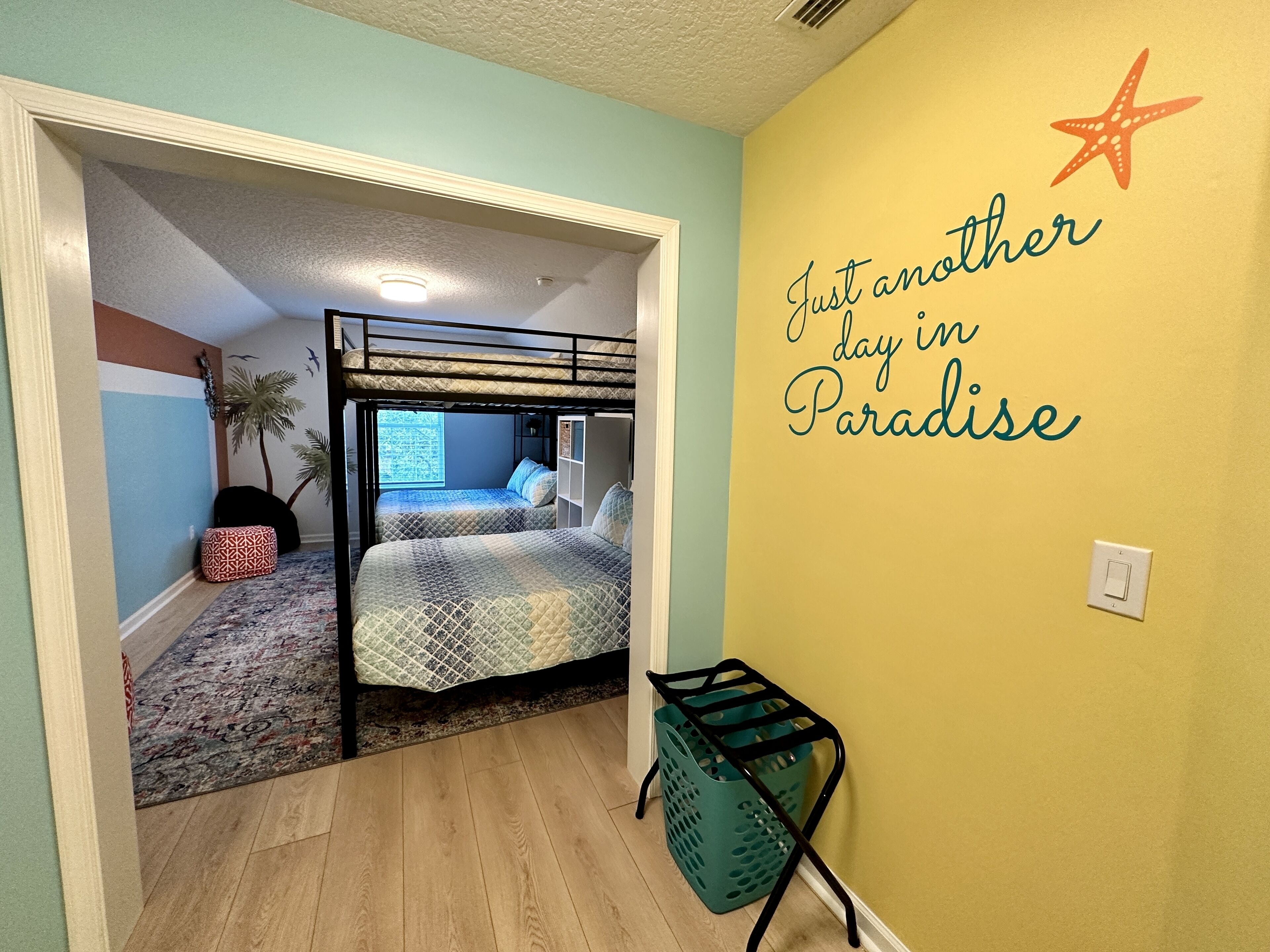 Une pièce avec un lit superposé, une petite table, un mur décoré d’une étoile et d’un texte, et une porte donnant sur une autre pièce.
