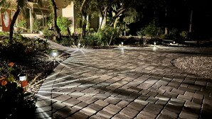 Un chemin pavé, bordé de part et d’autre par de la verdure et des arbres, et éclairé par des lampadaires régulièrement espacés.