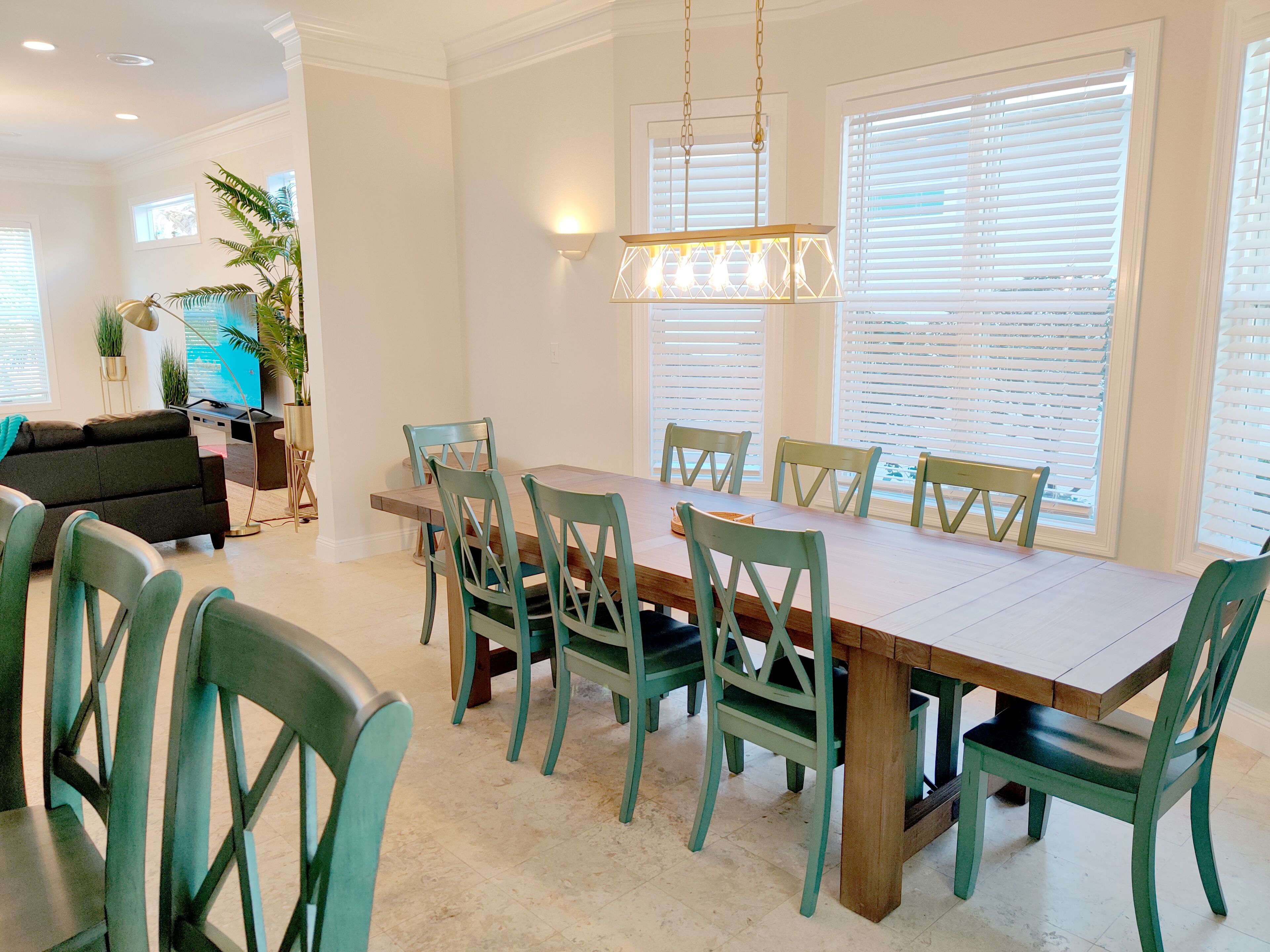 A dining area with a wooden table and teal chairs, surrounded by white walls and large windows with blinds.