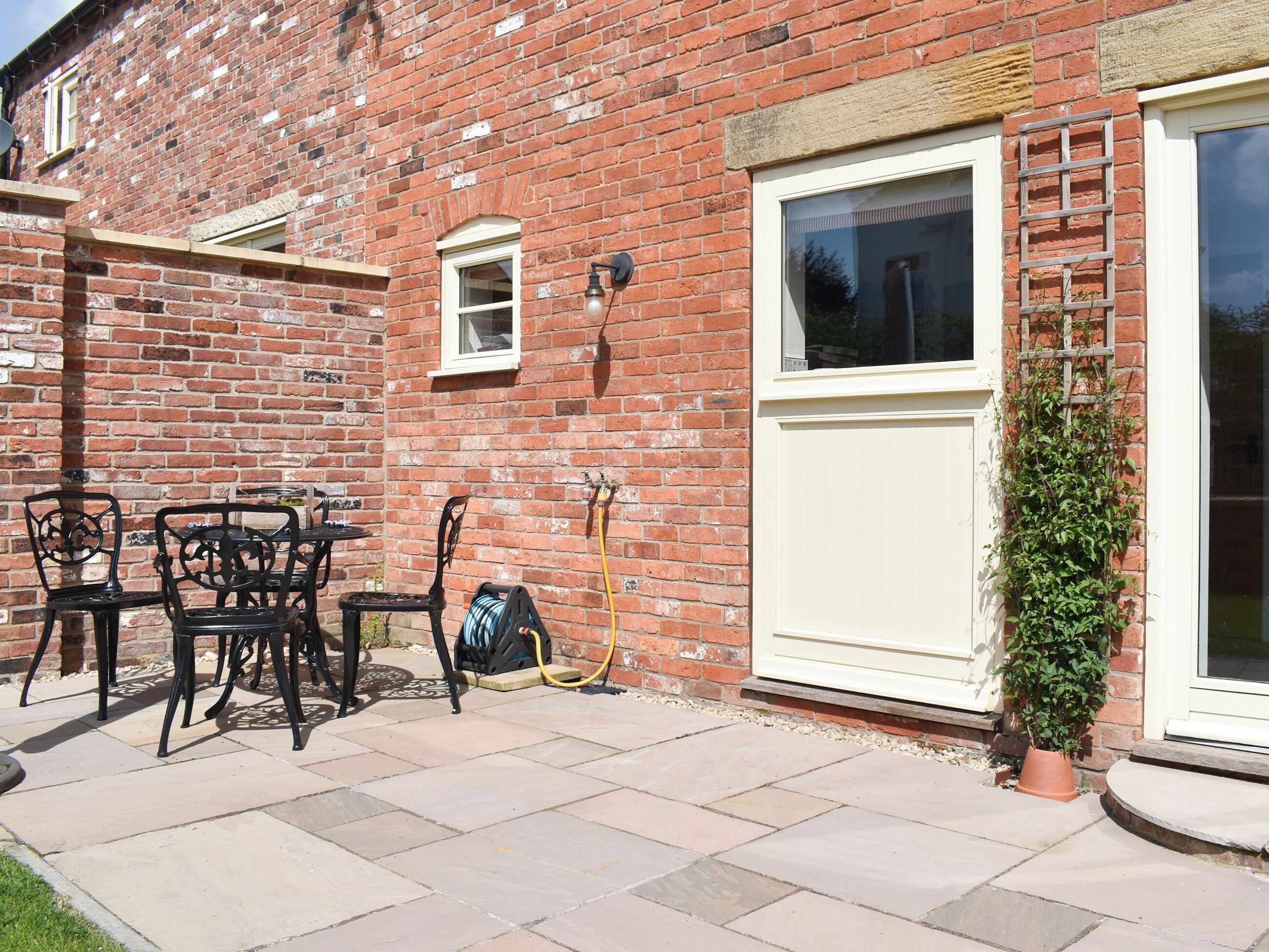 A brick building with a white door, a window, and a small window above the door. There is a garden hose and a black metal chair outside.