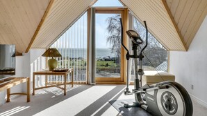 A spacious room with a large window offering a view of the sea, a ceiling fan, and a stationary exercise bike. - Gorgeous home in øster assels (Øster Assels)