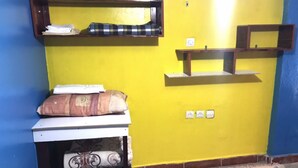A room with a yellow wall, a white table with a patterned cushion, and a shelf with folded items. - Furnished studio (El Ouatia)