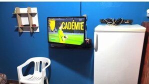 A room with a television mounted on the wall displaying a sports game, a white plastic chair, and a white refrigerator. - Furnished studio (El Ouatia)
