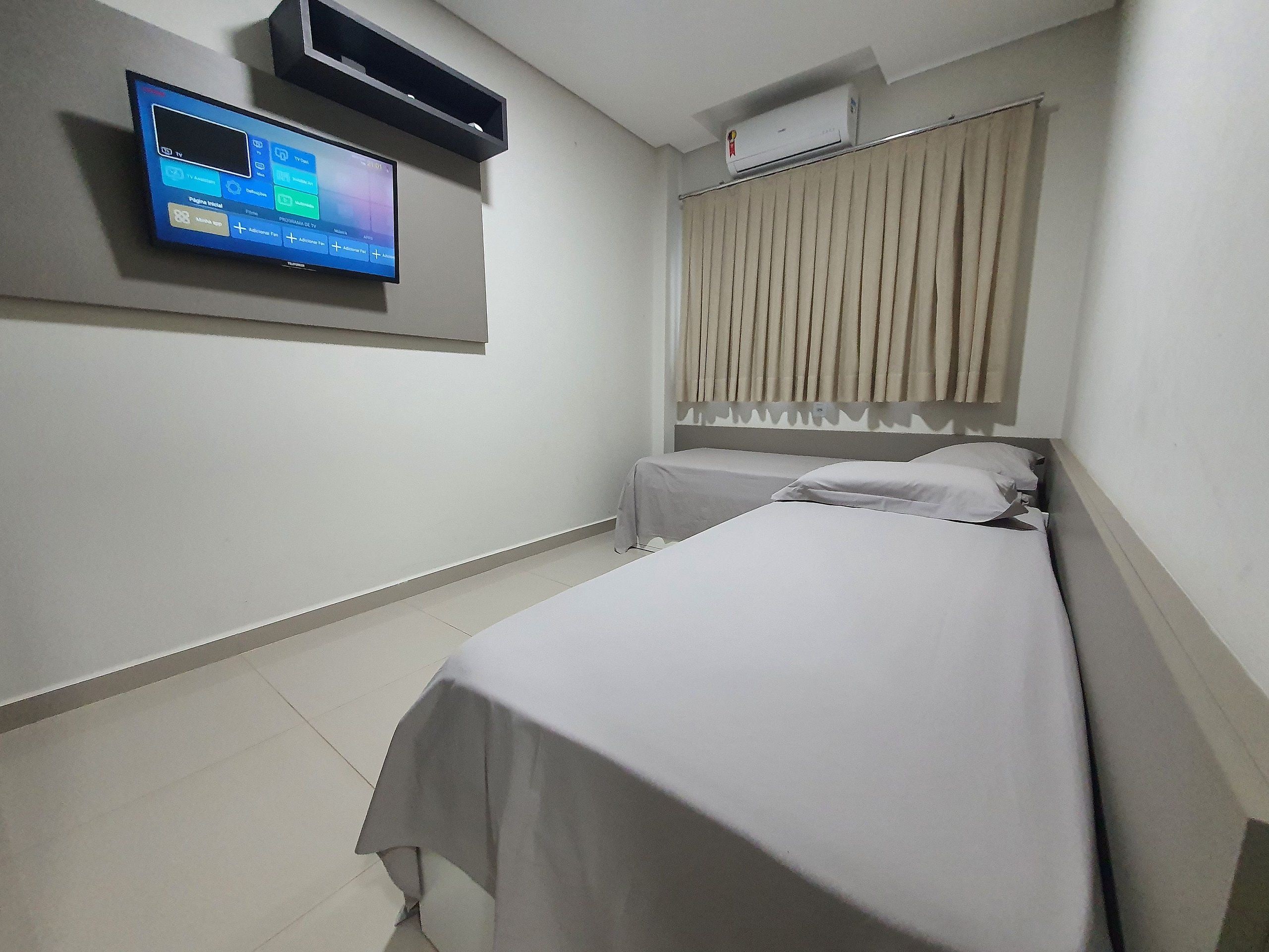 A hotel room with a bed, a TV mounted on the wall, and an air conditioning unit.