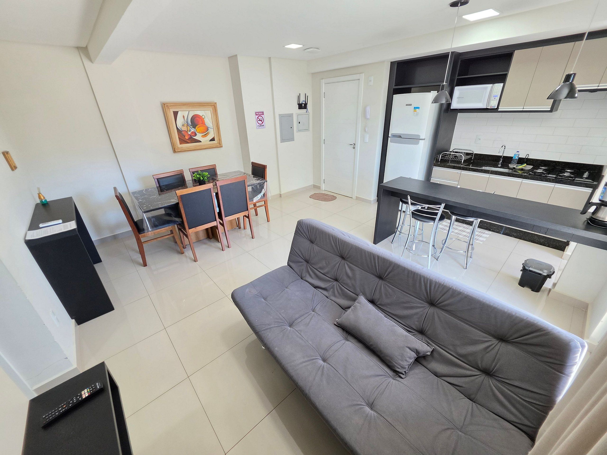 A modern apartment with a grey sofa, a dining area with a table and chairs, a kitchen with a microwave, and a painting on the wall.