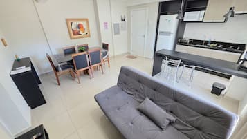 Eine moderne Wohnung mit einem grauen Sofa, einer Essecke mit Tisch und Stühlen, einer Küche mit einer Mikrowelle und einem Bild an der Wand.