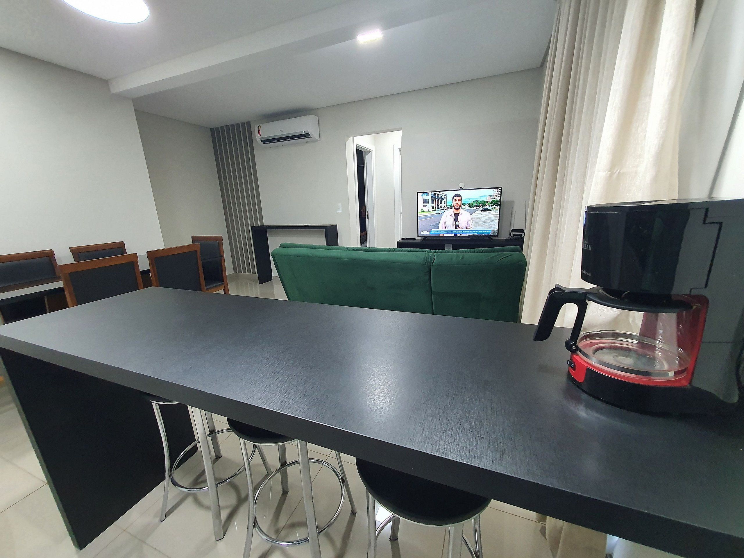 Cozinha moderna com bancada preta, máquina de café e televisão exibindo um noticiário.