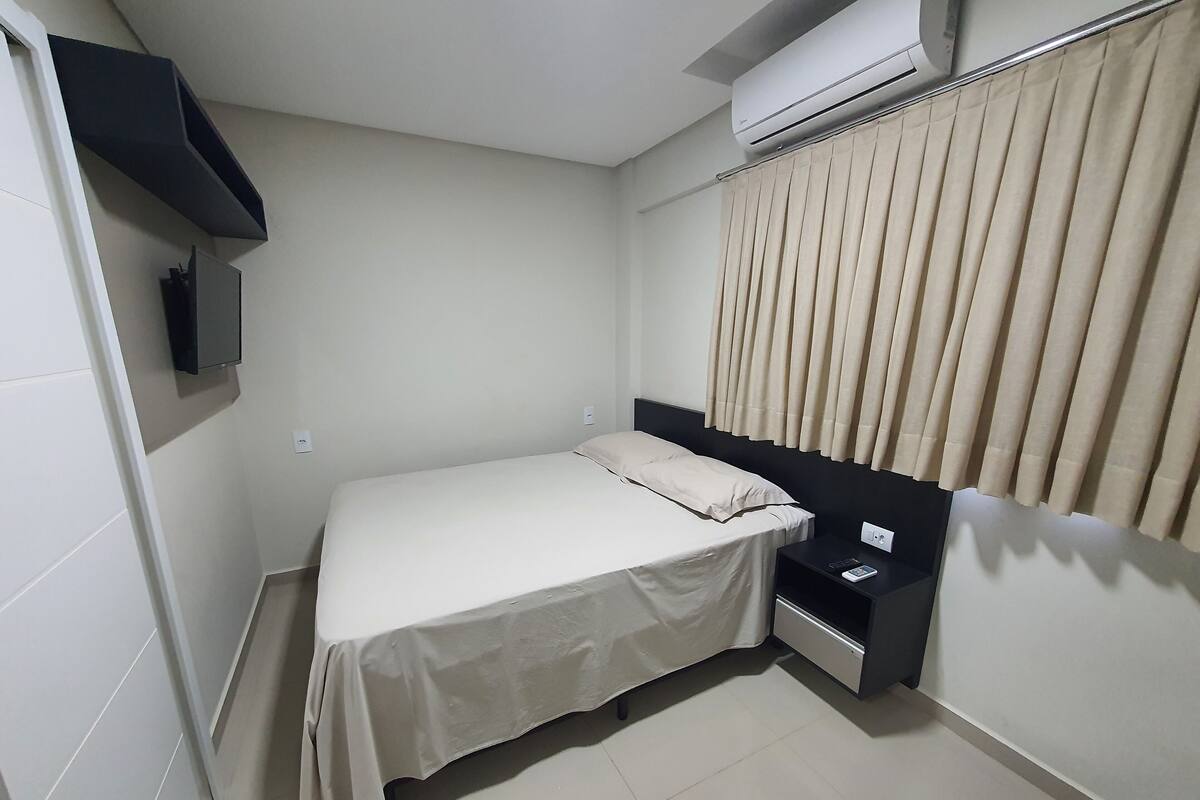 A small, clean room with a bed, bedside table, and a wall-mounted air conditioner.