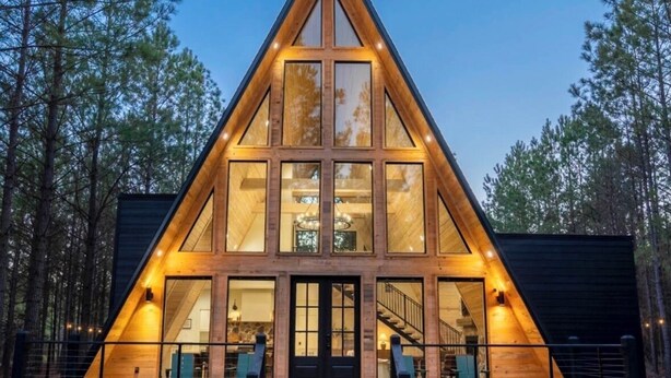 A modern, triangular wooden cabin with large windows, a stone foundation, and a staircase leading to the entrance.