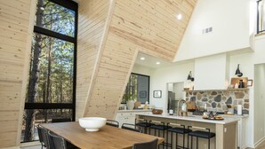 A modern kitchen with a central island, wooden beams, and a large window overlooking a forest.